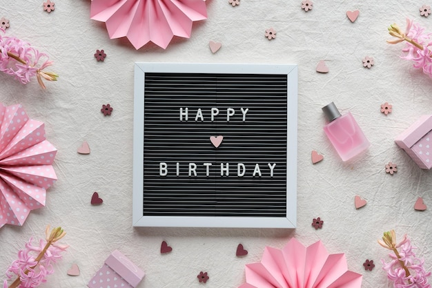 Testo buon compleanno sulla bacheca, letterboard. Piatto creativo, vista dall'alto su tessuto bianco crema. Decorazioni per feste, ventagli di carta, scatole regalo. Fiori di giacinto rosa naturale, bottiglia di vetro con profumo.