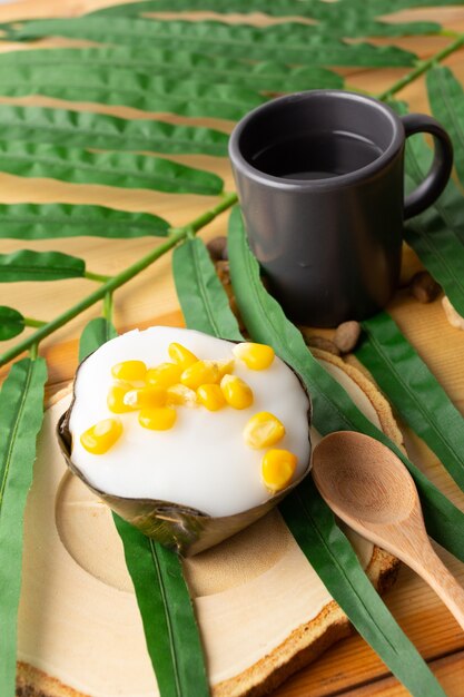 Thai Pudding Top in cocco con mais, dessert avvolto in foglie di banana. I thailandesi chiamano "Khanom Tako". Tako con mais posto su un piattino di legno e cucchiaio di legno, con tè in vetro nero.