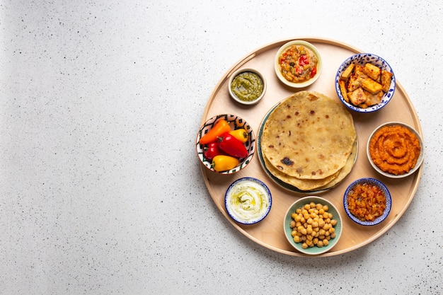 Thali indiano - selezione di vari piatti serviti su un piatto di legno rotondo, tavolo bianco. Meze vegetariano indiano assortito con pane chapati, paneer, dal, chutney in ciotole. Vista dall'alto, spazio per il testo