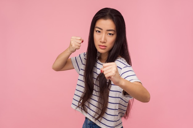 Ti prendo a pugni, Ritratto di ragazza arrabbiata con i capelli lunghi in maglietta a righe in piedi con gesto di boxe, pugni serrati pronta a combattere, minacciando di attaccare. girato in studio isolato su sfondo rosa