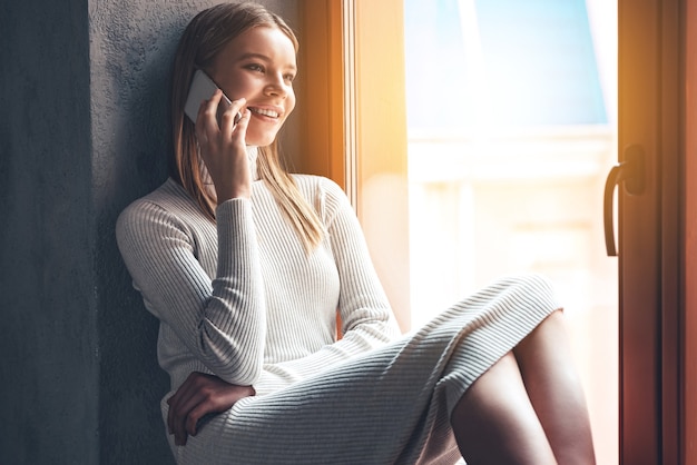 Ti sto aspettando! Bella giovane donna che parla al telefono cellulare con un sorriso mentre è seduta al davanzale della finestra