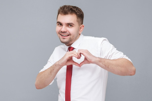 Ti voglio bene. Ritratto di giovane uomo felice bello in camicia bianca e cravatta in piedi con la forma di amore del cuore con le mani e guardando la fotocamera con la faccia felice. indoor isolato su sfondo grigio.