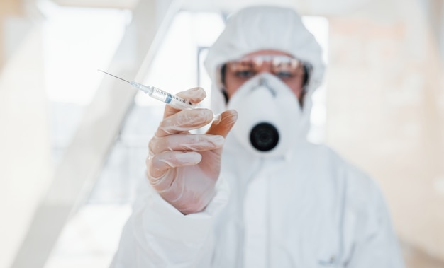 Tiene la siringa con la medicina. Scienziato medico femminile in camice da laboratorio, occhiali difensivi e maschera in piedi al chiuso