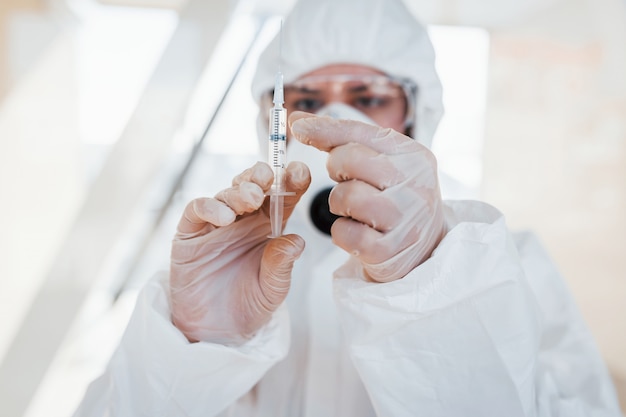 Tiene la siringa con la medicina. Scienziato medico femminile in camice da laboratorio, occhiali difensivi e maschera in piedi al chiuso