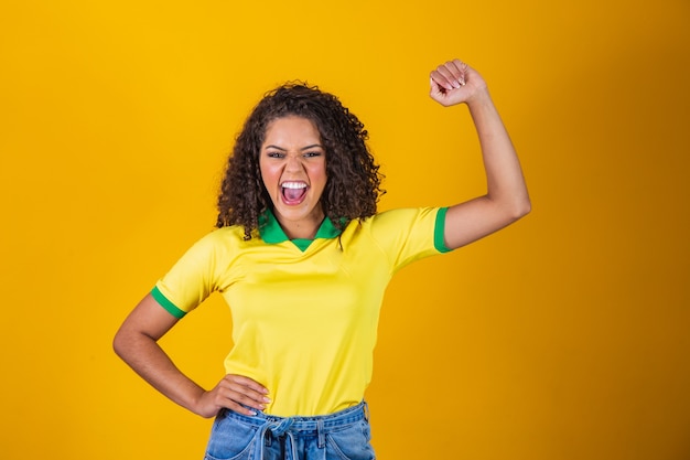 Tifoso del Brasile. Fan brasiliano della donna dei capelli ricci che celebra sul calcio, partita di calcio su fondo giallo. Colori del Brasile.