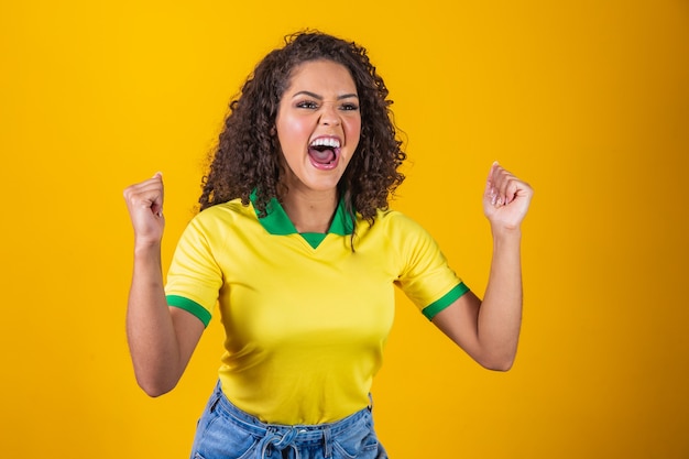 Tifoso del Brasile. Fan brasiliano della donna dei capelli ricci che celebra sul calcio, partita di calcio su fondo giallo. Colori del Brasile.