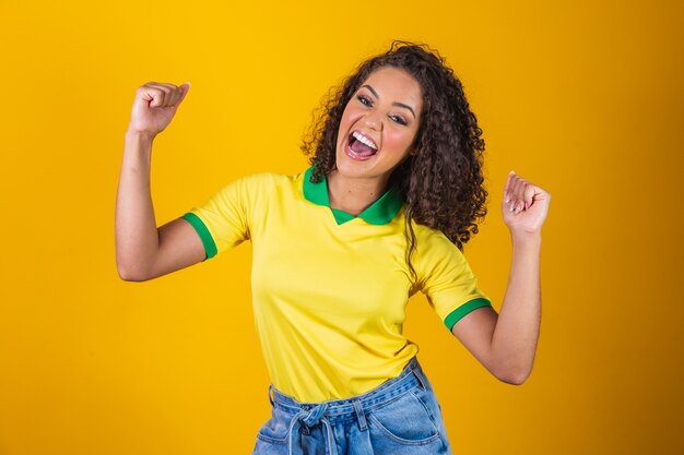 Tifoso del Brasile. Fan brasiliano della donna dei capelli ricci che celebra sul calcio, partita di calcio su fondo giallo. Colori del Brasile.