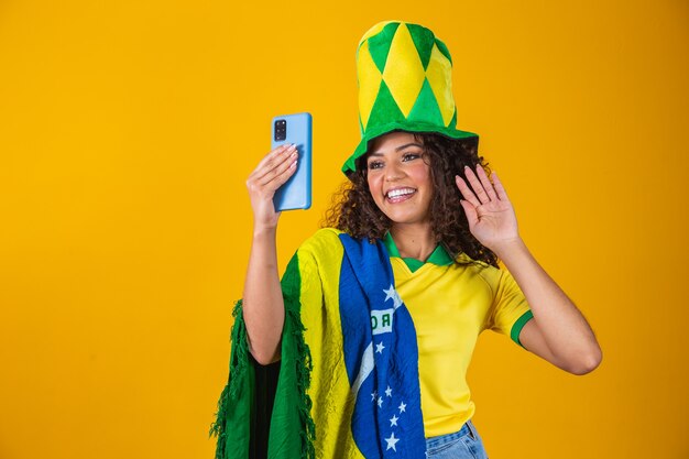 Tifoso del Brasile. Fan della donna brasiliana che celebra sul calcio, partita di calcio su sfondo giallo. Colori del Brasile. Smartphone per selfie.
