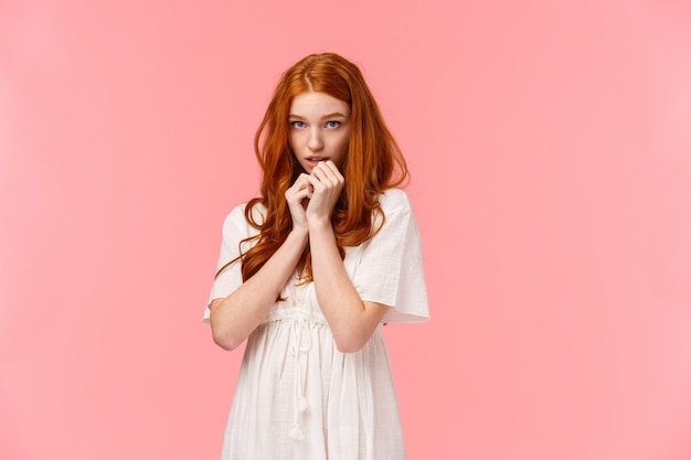 Timida kawaii, ragazza europea rossa in abito bianco, dall'aspetto timido e carino, arrossendo, guardando da sotto la fronte indecisa e timida, in piedi modesta sul muro rosa