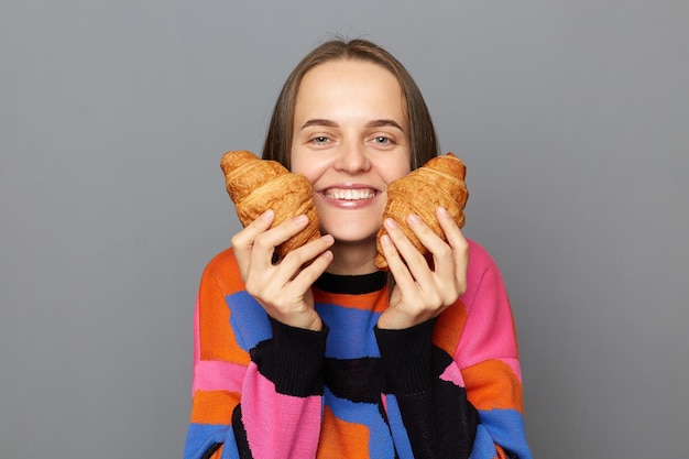 Tiro al coperto di felice felice giovane adulto caucasico donna che indossa un maglione in posa isolato su sfondo grigio sorridente alla telecamera dimostrando gustoso croissant