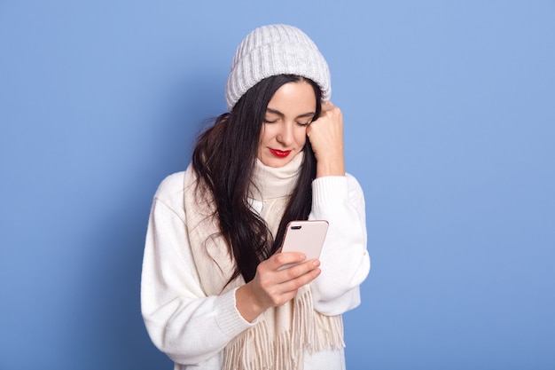 Tiro al coperto di sconvolto deluso ragazza bruna con gli occhi chiusi, in piedi isolato su uno spazio blu, tenendo lo smartphone in mano, tiene il pugno vicino al viso