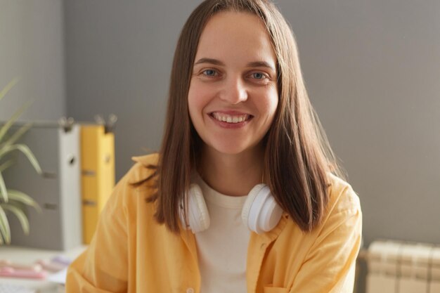 Tiro al coperto di sorridente felice giovane donna adulta con i capelli castani che indossa una camicia gialla e le cuffie sul collo seduto in ufficio e guardando la fotocamera con espressione felice