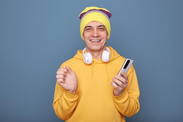 Tiro al coperto di un giovane uomo adulto soddisfatto e sorridente che indossa un berretto e una felpa con cappuccio gialla casual in piedi isolato su sfondo blu che tiene il telefono cellulare guardando la fotocamera con uno sguardo felice