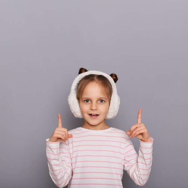 Tiro al coperto di una bambina stupita che indossa una camicia a righe e cuffie in pelliccia in piedi isolate su sfondo grigio che punta il dito indice verso l'alto nell'area pubblicitaria copia spazio per la promozione