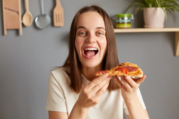 Tiro al coperto di una donna allegra felice che mangia un delizioso pezzo di pizza tenendo uno spuntino fast food guardando la fotocamera con il viso stupito che indossa una maglietta bianca casual in posa in cucina