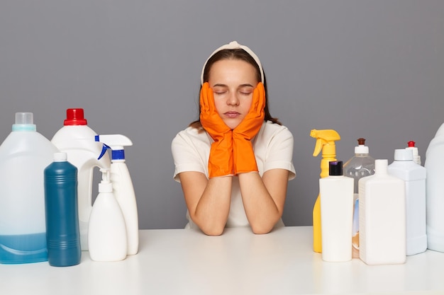 Tiro al coperto di una donna attraente esausta stanca in maglietta bianca e guanti arancioni in posa isolata su sfondo grigio seduta tra un sacco di detergenti con gli occhi chiusi riordinare tutta la casa si sente assonnata