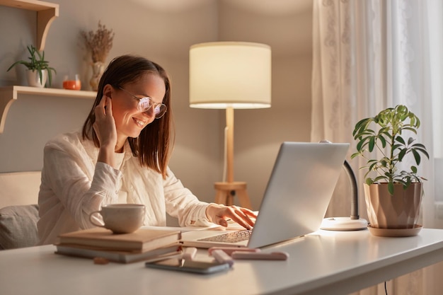 Tiro al coperto di una donna d'affari allegra e amichevole o di un libero professionista seduto in ufficio a casa utilizzando un computer portatile che guarda sorridente al display che lavora online la sera