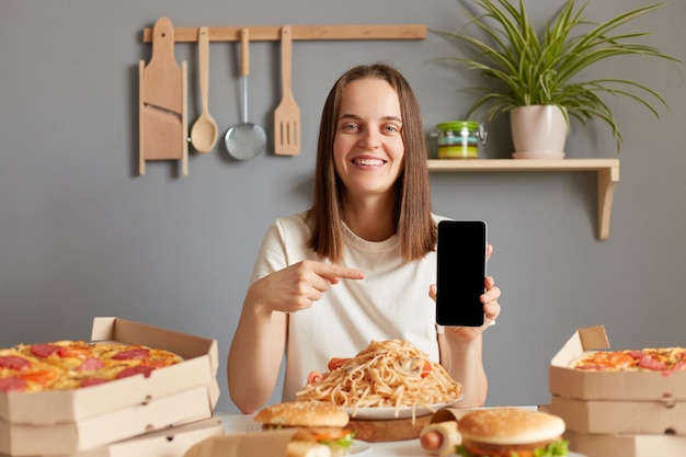 Tiro al coperto di una donna sorridente e gioiosa che indossa una maglietta bianca seduta al tavolo in cucina tra diversi piatti di fast food che mostra uno smartphone con display vuoto che raccomanda la consegna