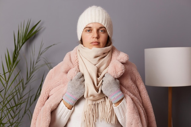 Tiro al coperto di una giovane donna caucasica adulta che indossa guanti da berretto e cappotto invernale guardando la fotocamera in piedi contro il muro grigio con lampada e casa dei fiori senza riscaldamento