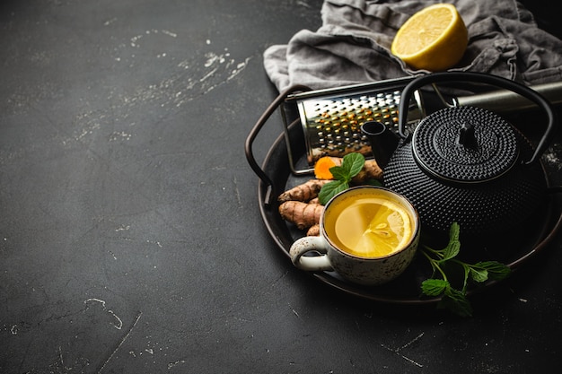 Tisana calda e salutare alla curcuma per rafforzare il sistema immunitario. Ingredienti per il tè detox in tazza, teiera nera, radice di curcuma fresca, limone, foglie di menta, grattugia su sfondo nero, vista dall'alto, spazio copia