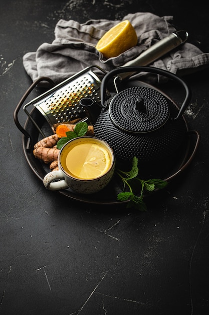 Tisana calda e salutare alla curcuma per rafforzare il sistema immunitario. Ingredienti per il tè detox in tazza, teiera nera, radice di curcuma fresca, limone, foglie di menta, grattugia su sfondo nero, vista dall'alto, spazio copia