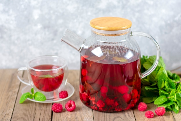 Tisana con frutti di bosco, lamponi, foglie di menta e fiori di ibisco in teiera di vetro e tazza sul tavolo di legno