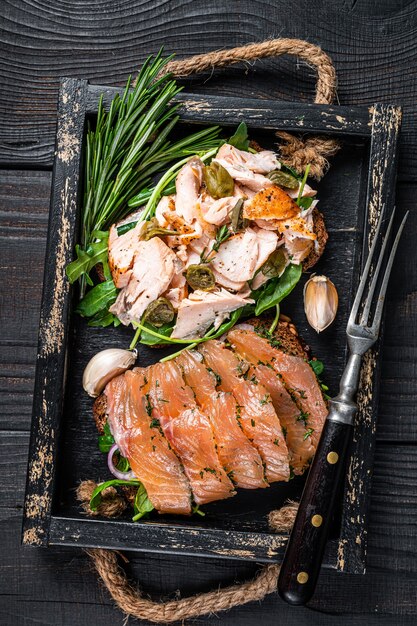 Toast con salmone affumicato caldo e freddo, rucola in vassoio di legno con erbe aromatiche. Fondo in legno nero. Vista dall'alto.