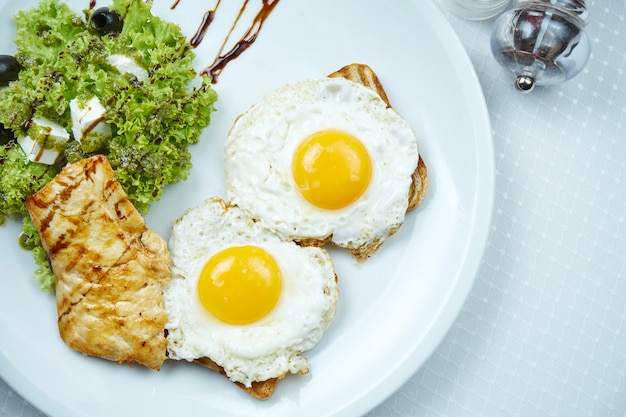 toast con uova fritte, filetto di pollo alla griglia, insalata con feta e olive sul piatto bianco.