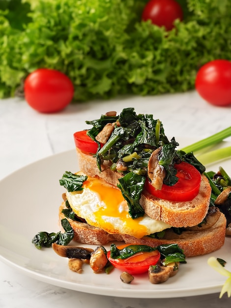 Toast con uova strapazzate su pane tostato con spinaci, funghi e pomodoro su un piatto grigio. Avvicinamento. Uovo fritto per colazione.