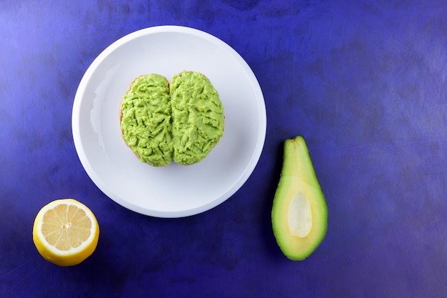 Toast e avocado con buccia verde e limone giallo su sfondo blu Toast salutari per colazione in primo piano con avocado schiacciato su un piatto bianco Vista dall'alto