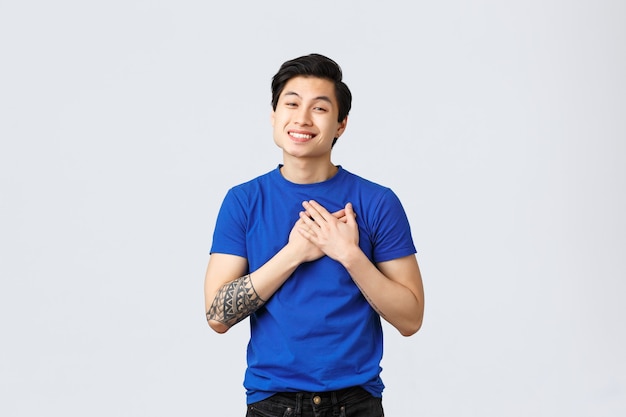 Toccato e grato bel maschio asiatico in maglietta blu con tatuaggi, toccando il cuore e sorridendo, apprezzando l'aiuto, essere contento o felice, ricevere complimenti commoventi, sfondo grigio