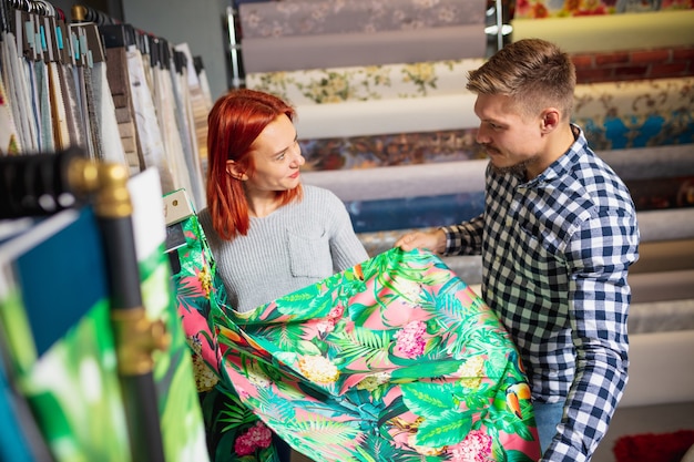 Tocco di stoffa. Coppia la scelta di tessuti nel negozio di decorazioni per la casa, negozio. Realizzazione di interior design per la casa durante la quarantena. Uomo e donna felici, la giovane famiglia sembra sognante, allegra scegliendo i materiali.