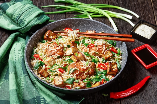 Tofu in agrodolce, cosce di pollo, noodles wok cinesi, cavolo nero, semi di sesamo, peperoncino rosso, cipolla verde, serviti su una padella con le bacchette su uno sfondo di legno scuro, vista dall'alto, primo piano