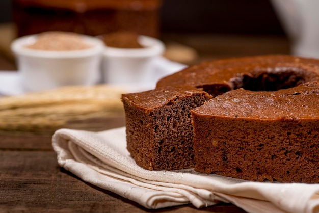 Torta al cioccolato e fetta su tessuto su legno con torta sullo sfondo con ramo di grano accanto.