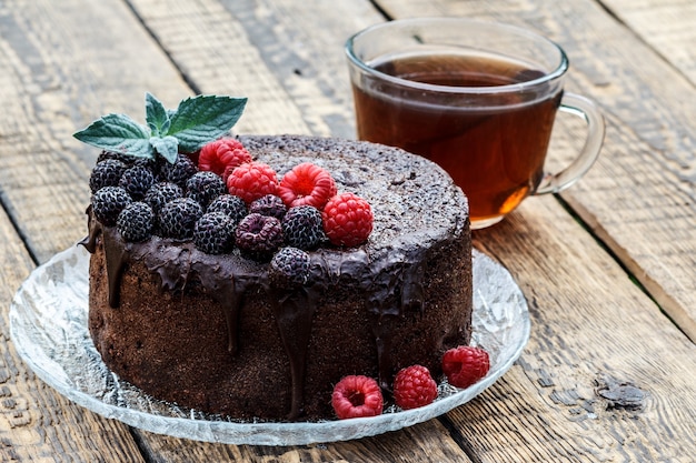 Torta al cioccolato fatta in casa decorata con foglie di menta, lamponi neri e rossi su lastra di vetro con tazza di tè su tavole di legno