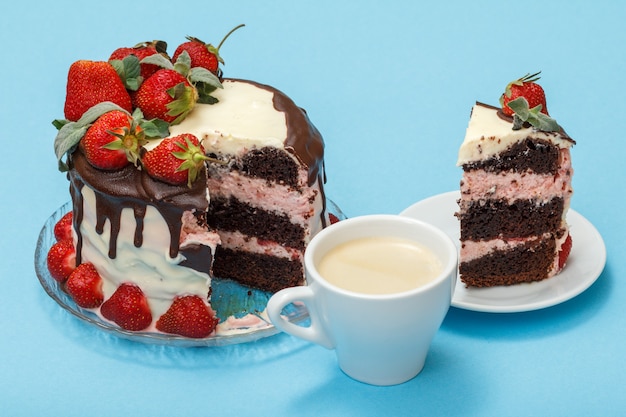 Torta al cioccolato fatta in casa decorata con fragole fresche e foglie di menta su lastra di vetro, tazza di caffè e fetta di torta su piattino bianco su sfondo blu