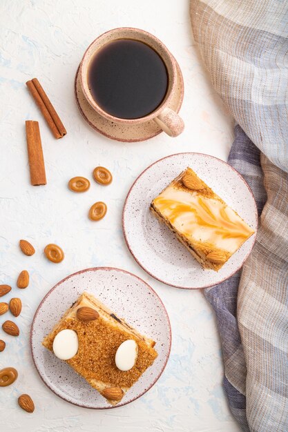 Torta al miele con crema di latte, caramello, mandorle e una tazza di caffè su fondo di cemento bianco e tessuto di lino. Vista dall'alto, piatto, primo piano.