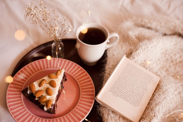 Torta con noci di mandorle su vassoio in legno con tazza di tè e libro aperto a letto. Buon giorno. Ora di colazione.