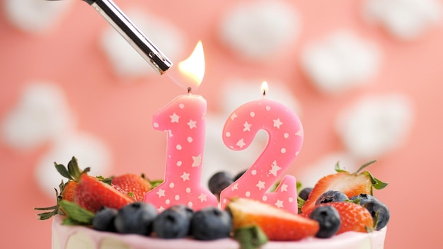 Torta di compleanno numero 12 candela rosa sulla bella torta con frutti di bosco e accendino con fuoco sullo sfondo di nuvole bianche e cielo rosa Vista ingrandita