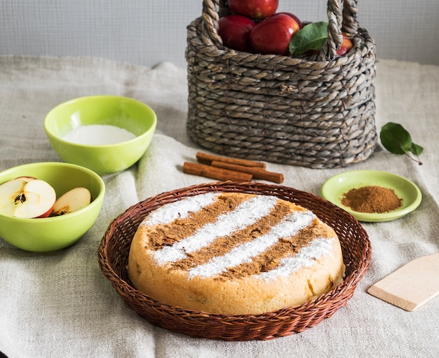 Torta di mele rotonda alla cannella fatta in casa in un piatto di vimini con ingredienti per cucinare in giro