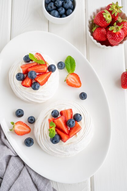 Torta di meringa Pavlova con panna montata e frutti di bosco freschi in cima su un piatto su fondo di legno bianco. Dolce estivo. Copia spazio, vista dall'alto.