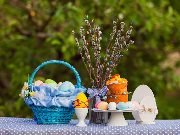 Torta di Pasqua fatta in casa su un supporto bianco con uova multicolori vicino Cesto blu splendidamente decorato con nastro Tavolo con decorazioni in stile pasquale e rami di salice in vaso di vetro