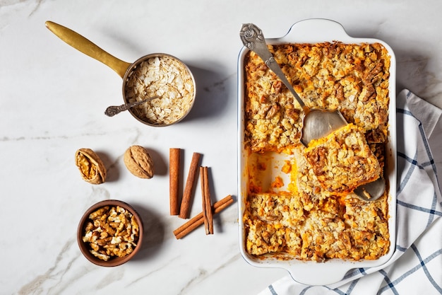 Torta di zucca con spezie tradizionali per torta di zucca: cannella, chiodi di garofano, noce moscata con noci e fiocchi d'avena in cima servita su una teglia con ingredienti su sfondo di marmo bianco, vista dall'alto