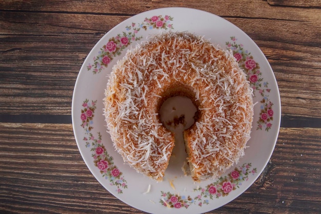 Torta dolce al gusto di cocco condita con scaglie di cocco con una fetta tagliata che mostra il delizioso impasto su un piatto decorato Utilizzando una tavola di legno e uno sfondo di legno Messa a fuoco selettiva Vista dall'alto