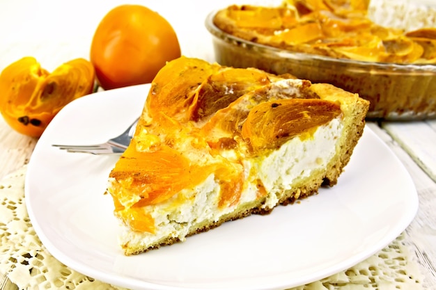 Torta dolce con cagliata e cachi, una forchetta nel piatto bianco su un tovagliolo di silicone, padella di vetro con torta su uno sfondo di assi di legno