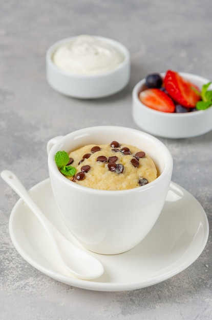 Torta in tazza con gocce di cioccolato, frutti di bosco freschi e panna su uno sfondo grigio cemento.