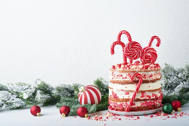 Torta natalizia a molti strati decorata con lecca-lecca coriandoli rossi e bianchi su supporto per torta bianca Palle di Natale rami di abete su tavolo di cemento bianco Messa a fuoco selettiva