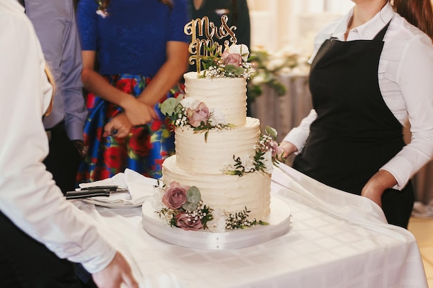 Torta nuziale moderna al ricevimento di nozze Camerieri che tirano fuori elegante torta nuziale bianca con decori floreali e topper mr mrs al banchetto di nozze nel ristorante Catering di lusso