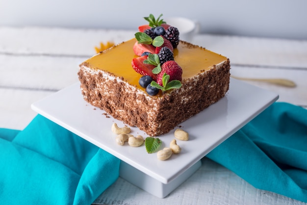 Torta quadrata fatta in casa decorata sopra gelatina gialla e frutti di bosco con menta su sfondo chiaro.