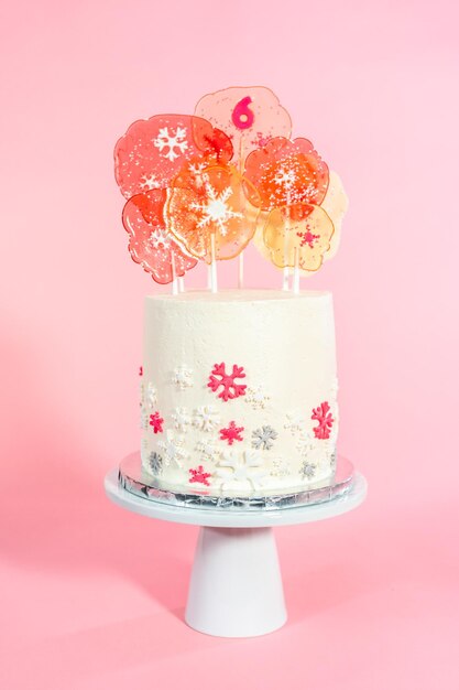 Torta rotonda alta con glassa di crema al burro italiana decorata con fiocchi di neve fondente e sormontata da grandi lecca-lecca rosa e bianchi su sfondo rosa.